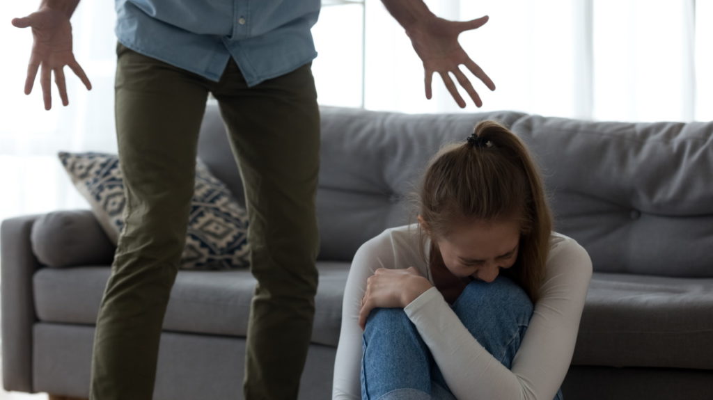 Unhappy frightened woman crying, sitting on floor, aggressive man shouting to scared wife, angry husband emotionally quarreling, arguing, emotional psychological abuse, domestic violence concept